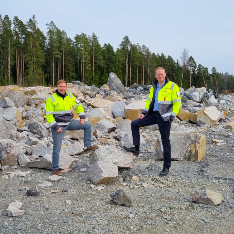 Two men in high-visibility jackets stand on a construction site, each with one foot on a large rock, looking directly at the camera.