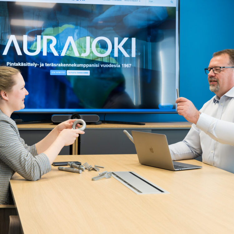 A woman and a man are sitting at a conference table, discussing small steel products in their hands. In the background, there is a screen displaying "Aurajoki - Your Surface Treatment and Steel Structure Partner Since 1967".