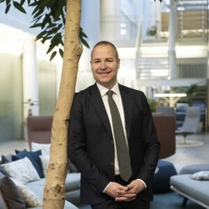 Pukuun pukeutunut mies nojaa huonekasvipuuhun, katsoo kameraan ja hymyilee.