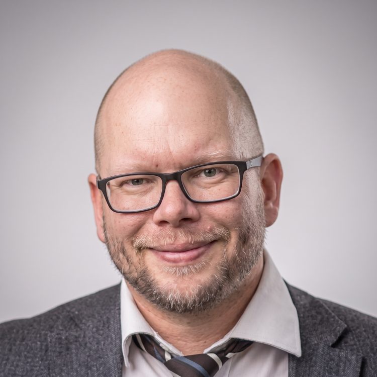 Mies katsoo kameraan kauluspaidassa ja löysytetyssä solmiossa ja hymyilee.